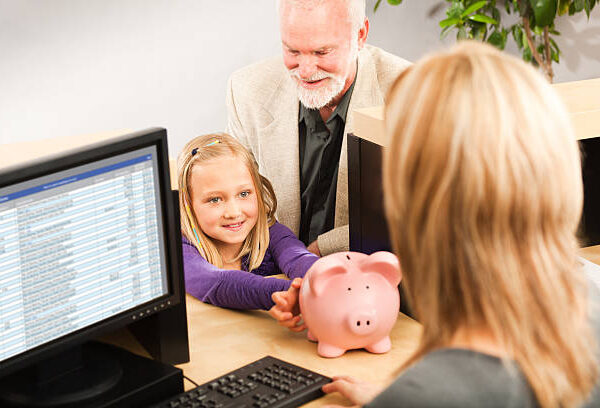 A cheerful grandparent helping a smiling young girl grandchild with her piggy bank savings. Opening a children bank account with the bank teller in a retail bank counter. Photographed indoors in horizontal format.