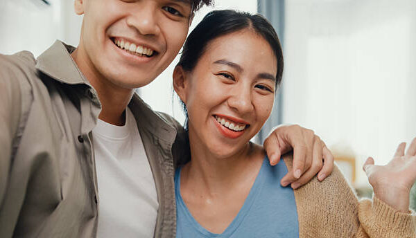 Young happy Asian sweet couple video call look at camera smiling so proud and show off new house to their friends in living room. Husband wife move new home, Apartment Rent And Ownership Concept.